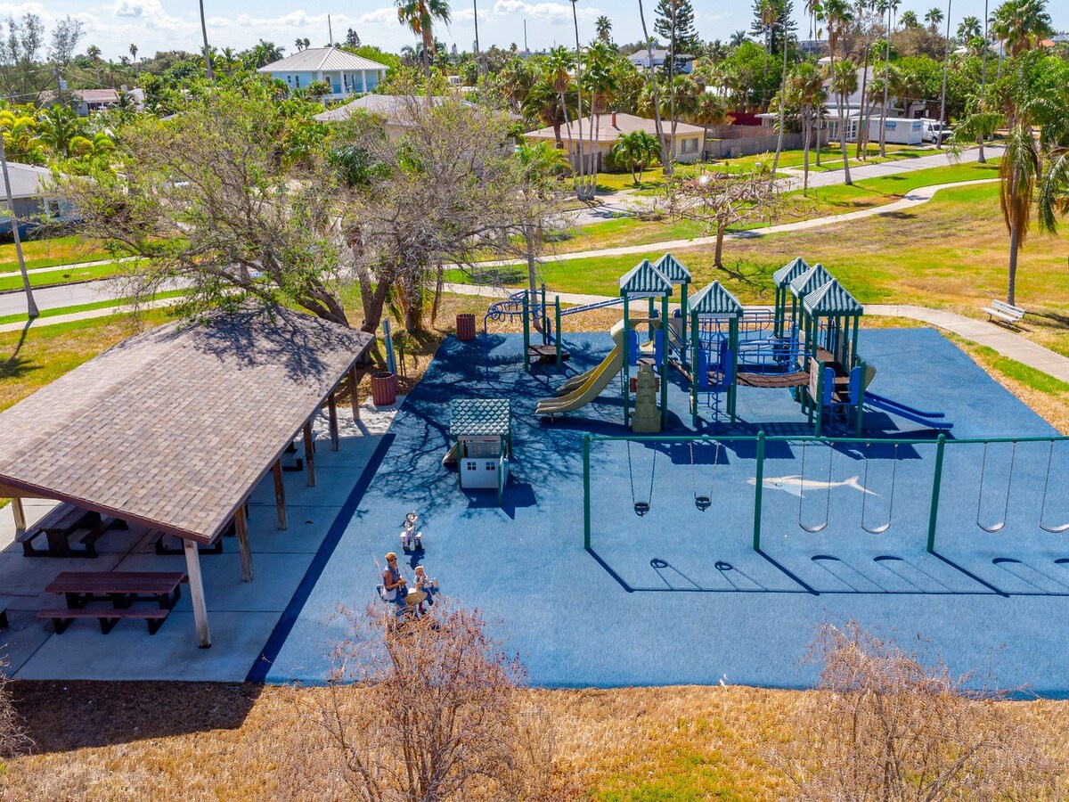 A colorful playground with swings, slides, and shaded picnic area sits within a residential neighborhood surrounded by palm trees and green spaces.
