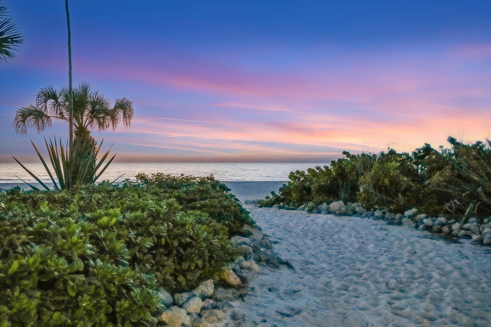 A sandy path winds through tropical greenery toward the ocean as the sky glows pink and gold at sunset, creating a tranquil coastal scene near the property.