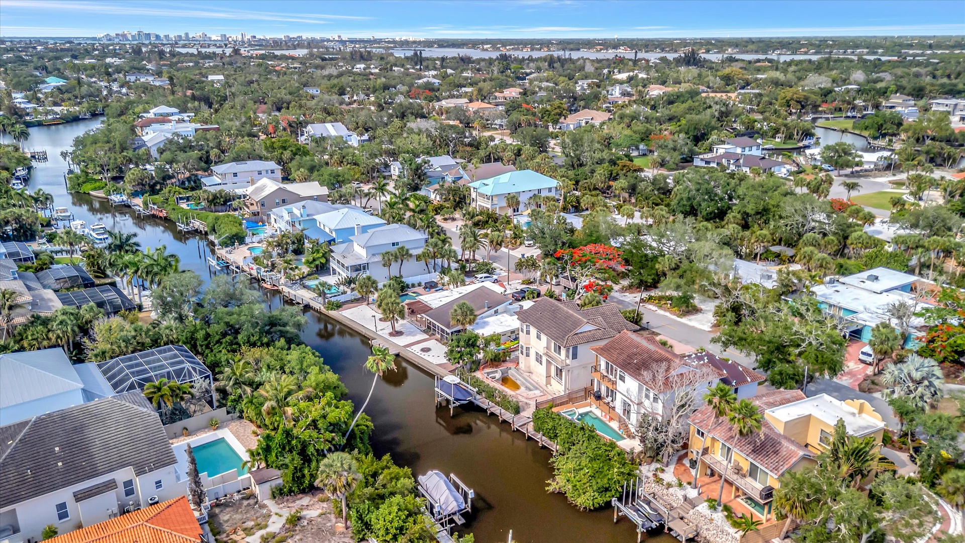 Aerial view of a waterfront neighborhood featuring canal-side homes with private docks, lush greenery, and proximity to urban amenities on the horizon.