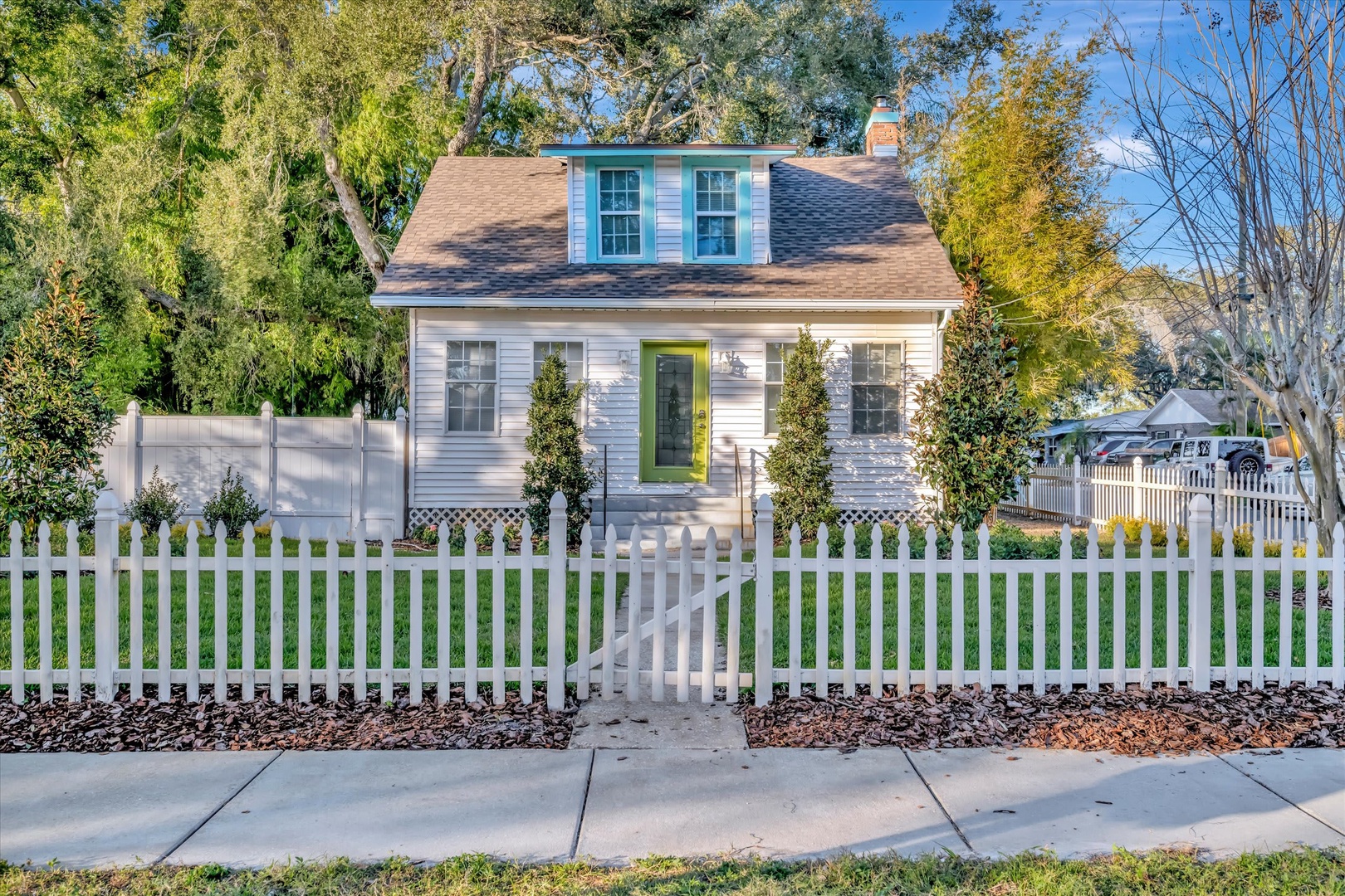 A charming cottage with a cheerful green door welcomes you from a quiet, tree-lined residential street. The classic white picket fence and manicured lawn create a cozy, storybook feel in this peaceful neighborhood setting.