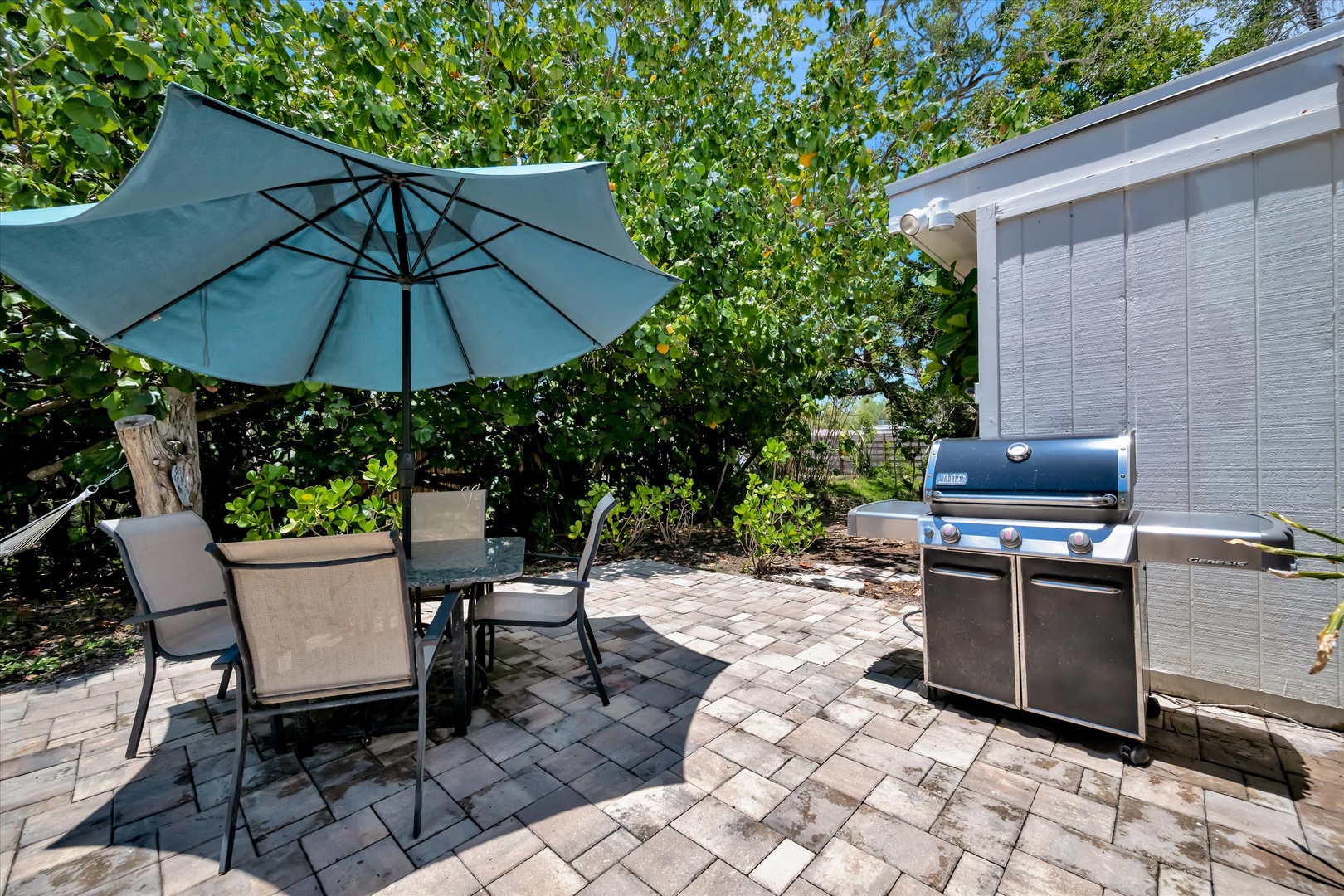 Your private patio beckons with lush greenery overhead, shaded dining under an umbrella, and a sleek grill ready for meals under the open sky.
