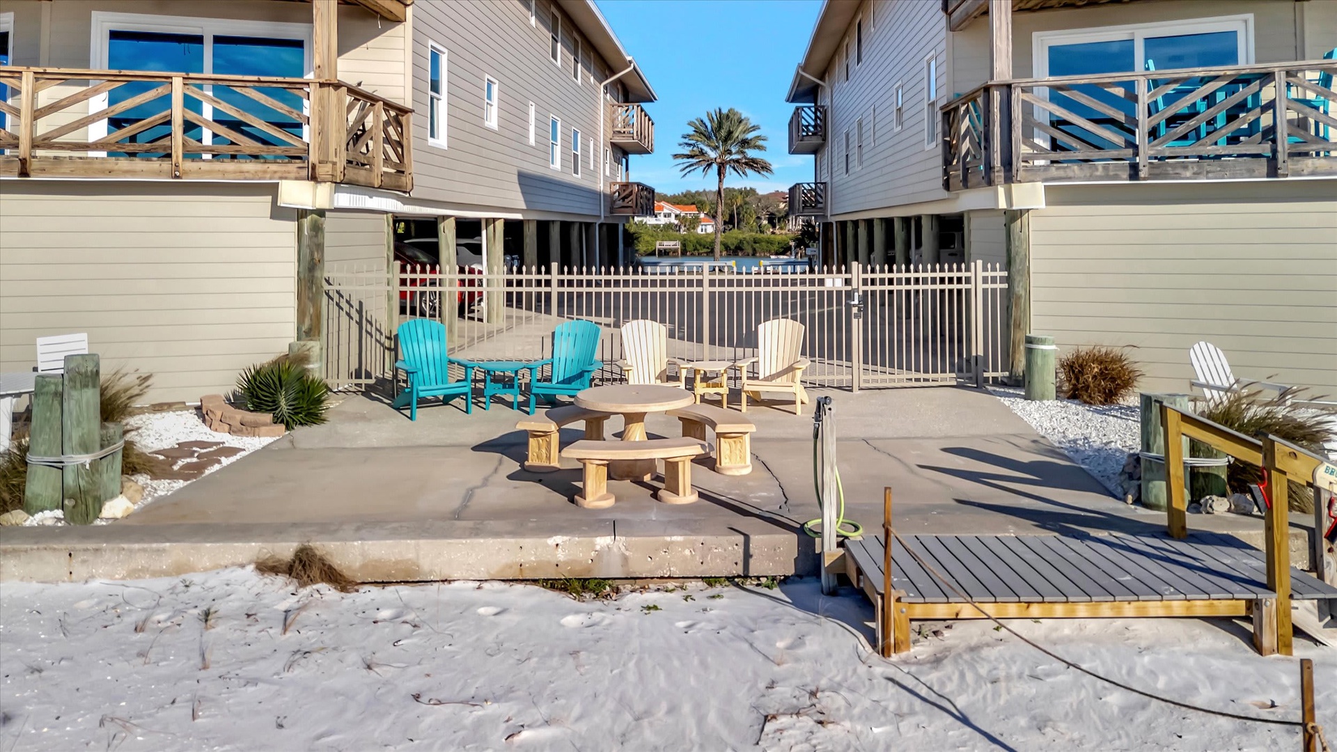 This beachfront property features direct sand access, a communal fire pit, colorful Adirondack chairs, and coastal boardwalk seating positioned between residential buildings.