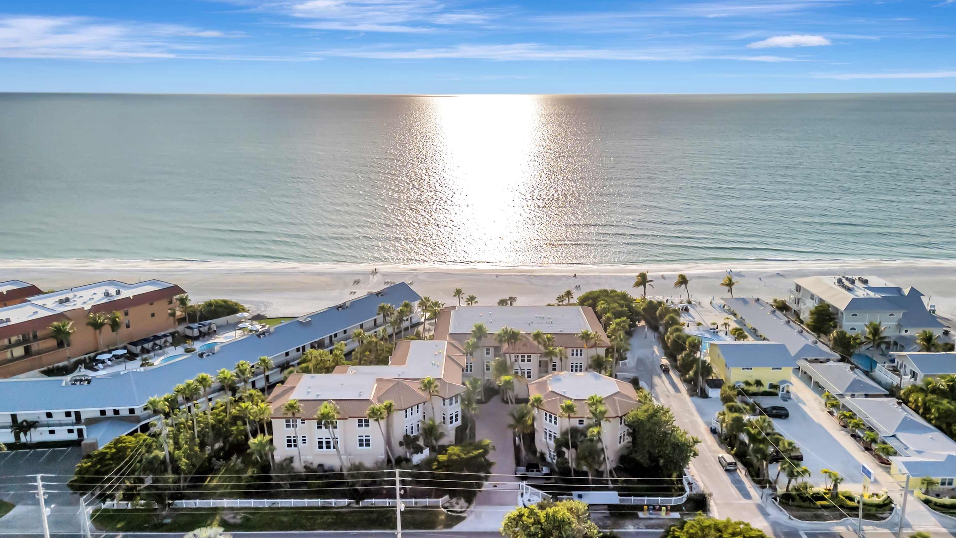 A pristine beachfront community stretches along the shore, framed by palm-lined streets and gentle coastal waters reflecting the bright sunlight overhead.