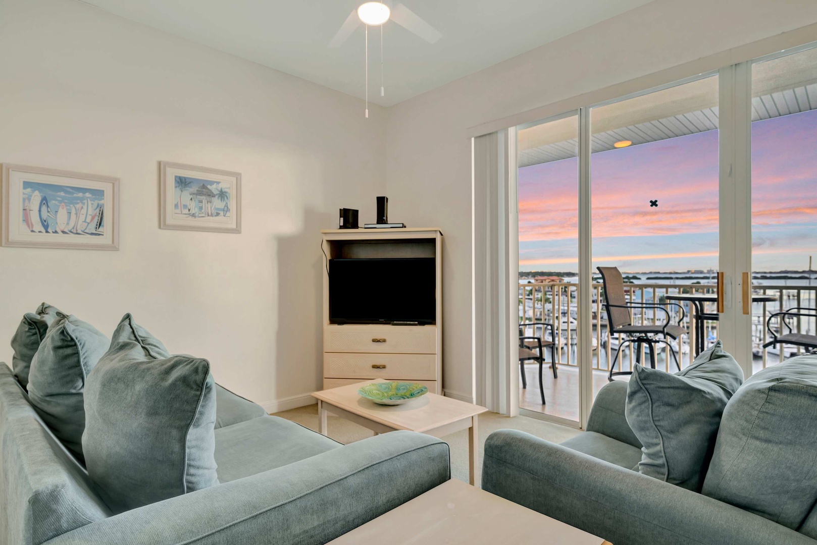 Step into a bright, serene living space where plush sofas invite you to unwind. Through the balcony doors, watch the sky paint itself in breathtaking sunset hues as evening settles over the water.