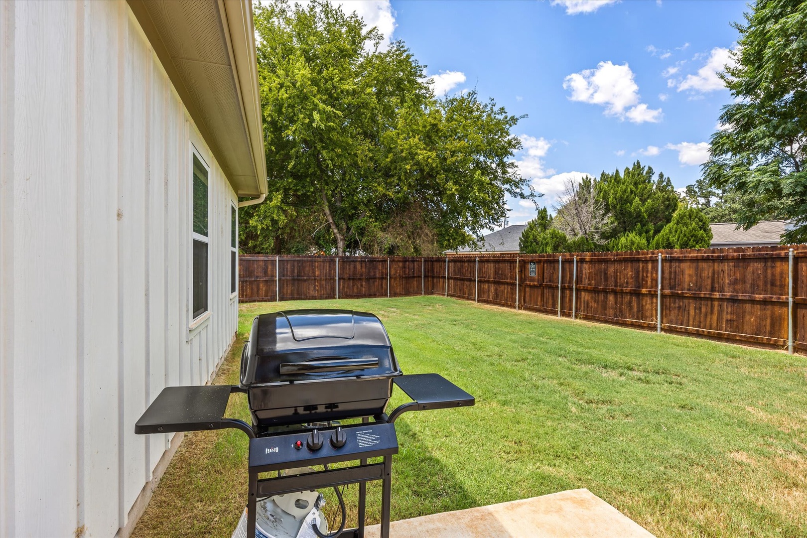 A private, fenced backyard with lush green lawn and mature trees. The outdoor grill awaits your summer cookouts under open skies.