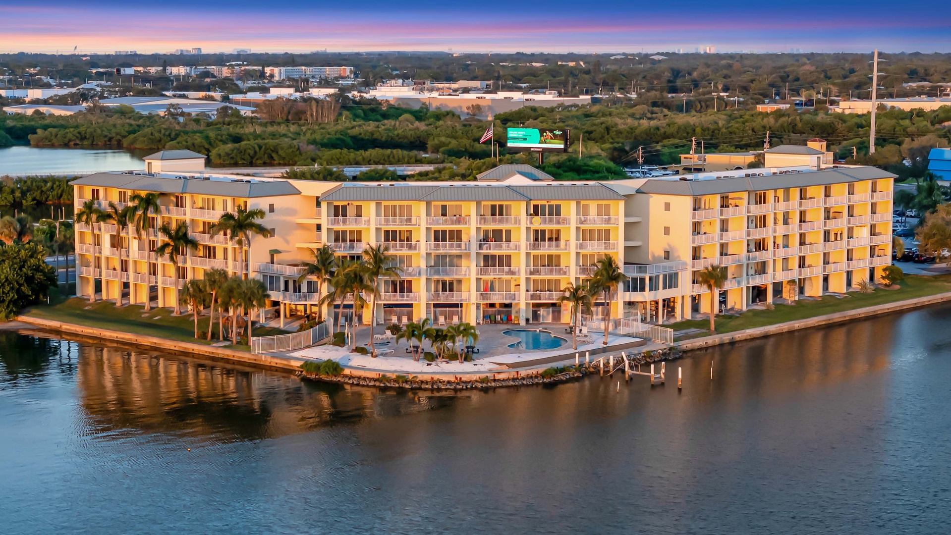 Aerial view of a waterfront resort property illuminated during golden hour, nestled along a calm waterway with tropical landscaping and urban surroundings.
