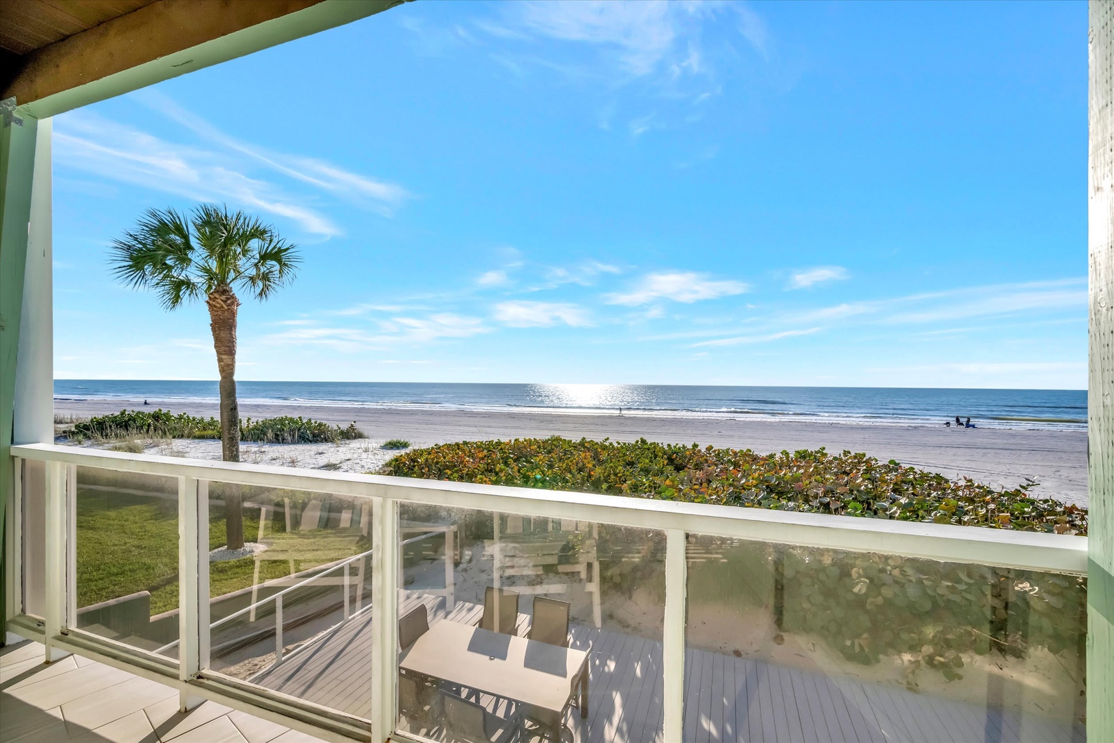 Beachfront property offering direct views across a sandy beach, coastal palms, and the ocean beyond. A white railing encloses the covered porch area.