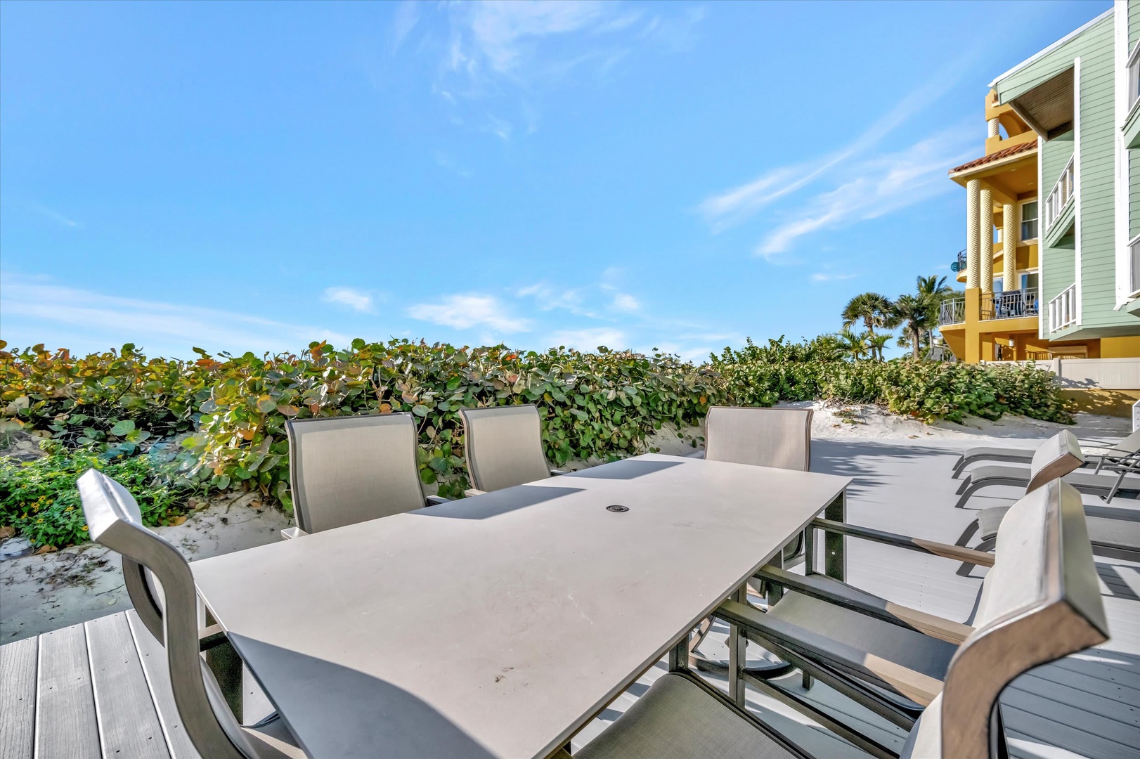 Beachfront outdoor dining area with a spacious table and chairs set against lush coastal foliage and sandy shores, with colorful vacation homes nearby.