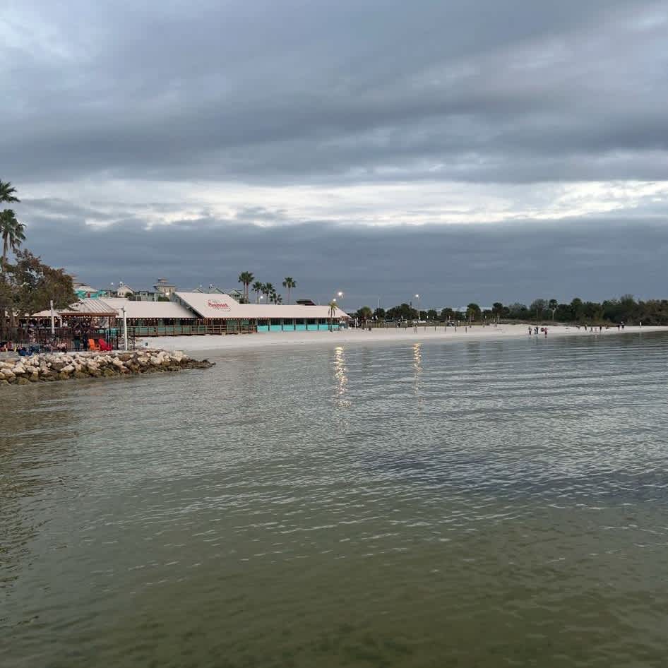 A calm beach area with gentle waters and a restaurant nearby, framed by palm trees and an expansive cloudy sky—a peaceful retreat in a coastal setting.