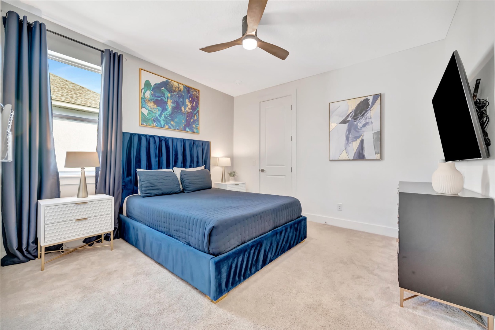 Relax in your serene bedroom featuring a luxurious velvet bed, modern ceiling fan, and curated artwork. Natural light fills the space, while the mounted TV and plush carpet ensure your comfort.