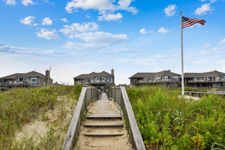 A boardwalk leads through coastal dunes to a row of beachfront vacation properties under a bright, cloud-dotted sky, offering easy access to the shore.