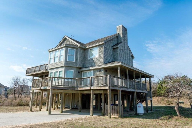 A coastal vacation home elevated on stilts with wraparound decks at each level, offering expansive views and classic beach architecture. Situated on an open lot with natural dune grasses.