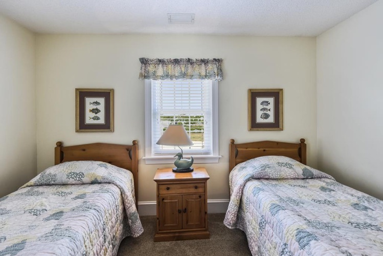 Your cozy twin bedroom awaits, with matching beds dressed in coastal quilts, warm wood furnishings, and natural light spilling through the window to brighten your peaceful retreat.