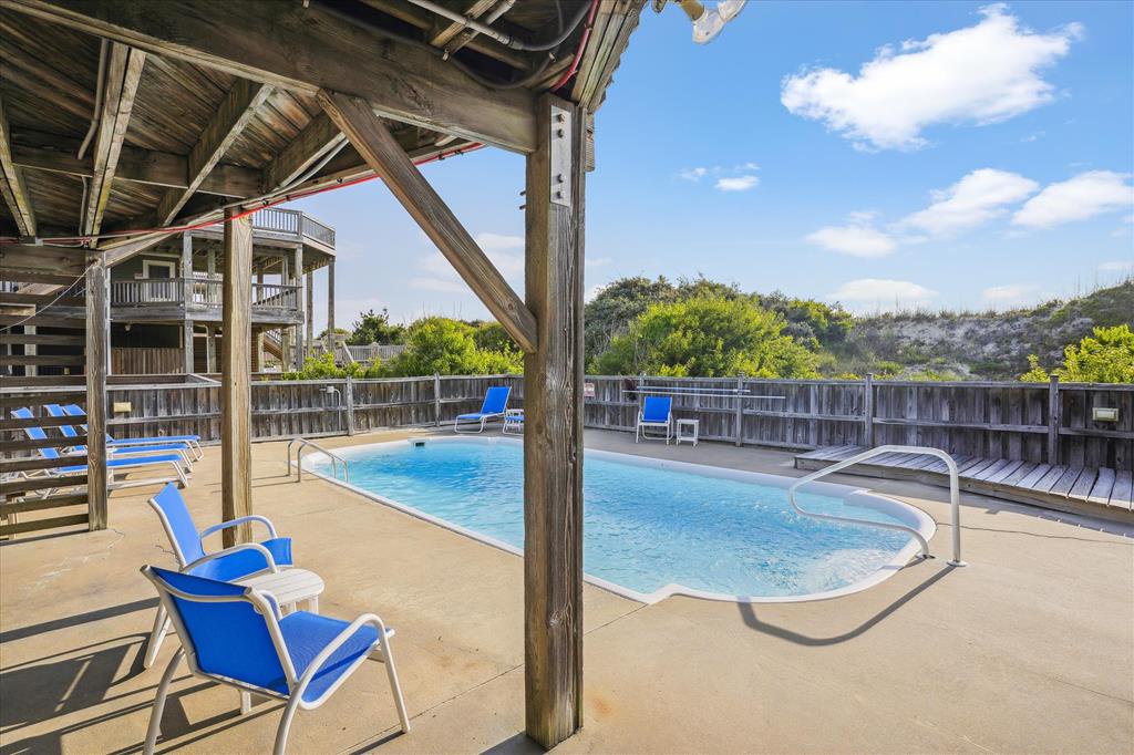 A serene outdoor pool area featuring two inviting pools surrounded by wooden decking and lounge chairs, set against a backdrop of lush greenery and blue skies.