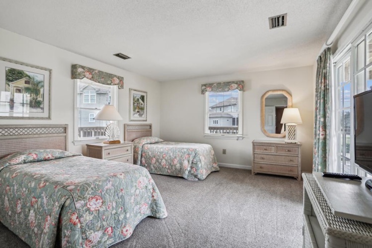 Step into your airy, sunlit bedroom with two plush beds dressed in floral linens, gentle natural light pouring through multiple windows, and a peaceful retreat waiting for your seaside escape.