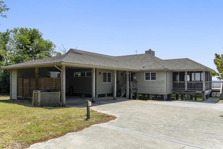 A single-story vacation home features covered porches and parking, surrounded by green lawn and mature trees under a clear blue sky.