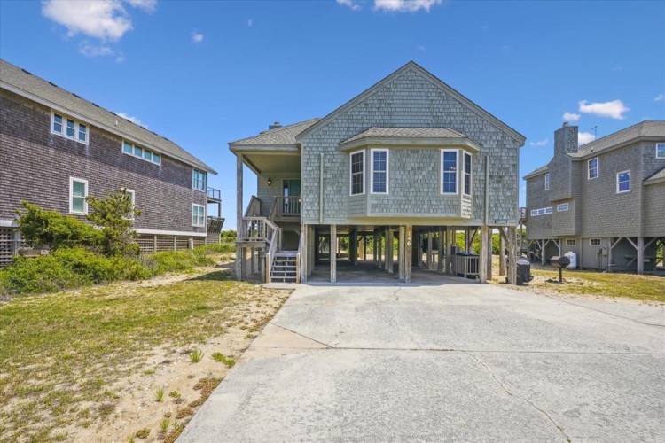 A charming coastal home features classic shingle siding and elevated architecture in a peaceful beachside neighborhood, with convenient parking and easy access to sandy shores nearby.