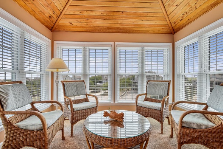 Relax in this bright sunroom with wicker seating, wooden vaulted ceilings, and panoramic windows framing serene outdoor views—a peaceful space to unwind any time of day.