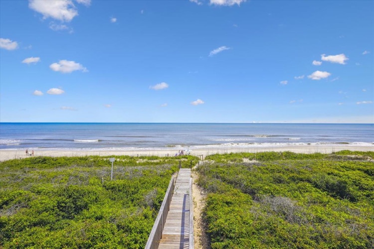 A scenic boardwalk leads through lush coastal vegetation to a pristine sandy beach, with gentle waves rolling in under a bright blue sky dotted with soft clouds.