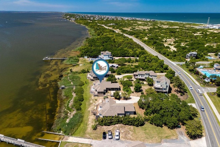 Aerial view captures a waterfront property nestled between calm bay waters and coastal landscape, with direct access and proximity to beach areas along a scenic coastal road.