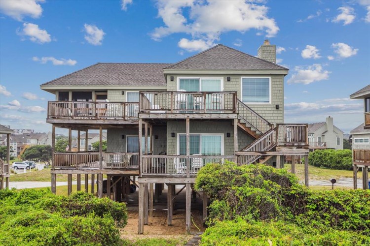 Coastal vacation home featuring multiple levels of decks and elevated stilts, nestled in a residential beachside neighborhood with lush landscaping and easy beach access nearby.