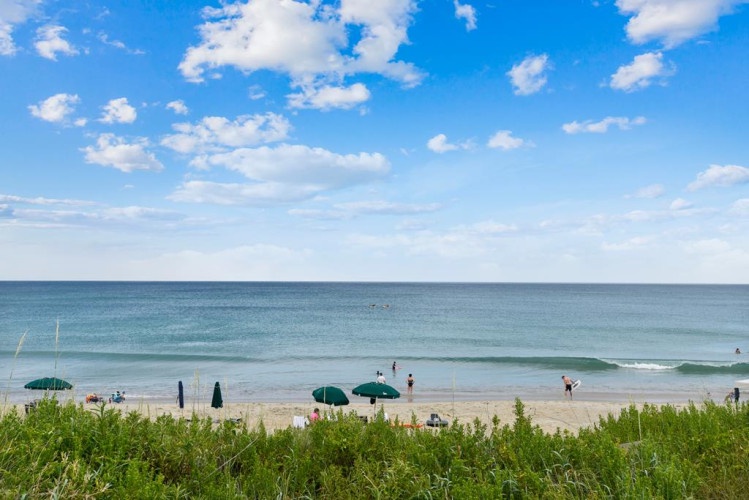 A pristine beach scene with gentle waves rolling ashore, umbrellas dotting the sand, and natural dune grasses swaying in the coastal breeze under a beautiful blue sky.