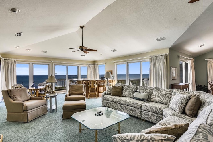Imagine lounging in this spacious living room with sweeping ocean views through wall-to-wall windows. Your toes sink into soft carpet as sea breezes drift through. Unwind on plush seating, gather at the dining table, and soak in endless blue horizons.