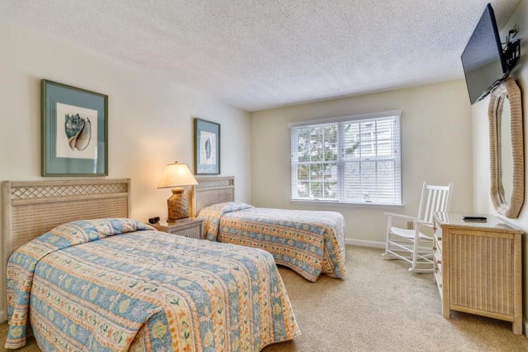 Your coastal-inspired guest room features twin beds with woven headboards, beach-themed art, and a rocking chair by the window—perfect for relaxing after sun-filled days.