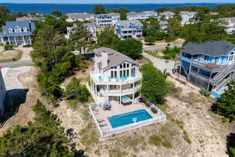 Aerial view of a three-story coastal vacation home with private pool, multiple decks, and proximity to neighboring beach houses and waterfront access.