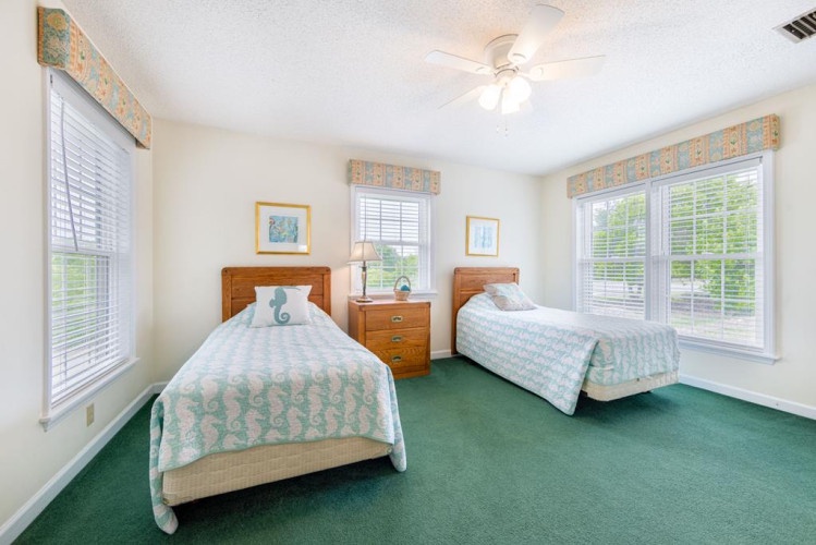 Your bright, peaceful twin bedroom features plush carpeting, ample natural light from generous windows, and coastal-inspired bedding that invites restful nights. Ceiling fans and wood furnishings add warmth and comfort to your home away from home.