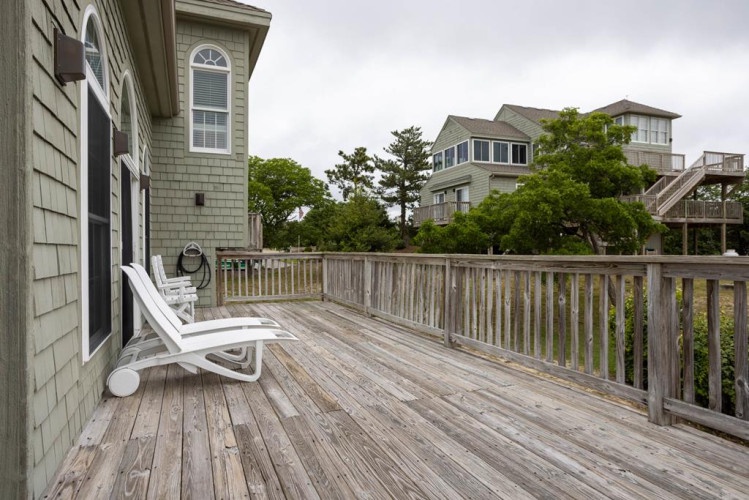 A spacious wooden deck with comfortable lounge seating, surrounded by coastal greenery and neighboring beach homes, offering a perfect outdoor retreat for your seaside getaway.