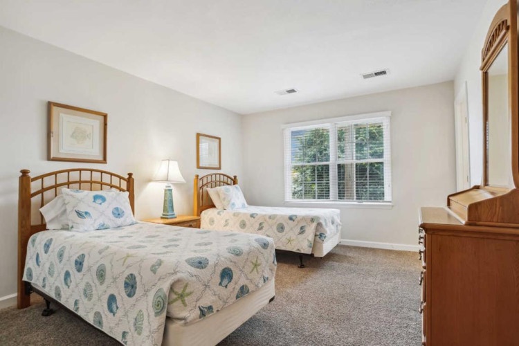 Relax in your bright, peaceful bedroom featuring two cozy beds, warm wood furnishings, and natural light streaming through the window to start your day refreshed.