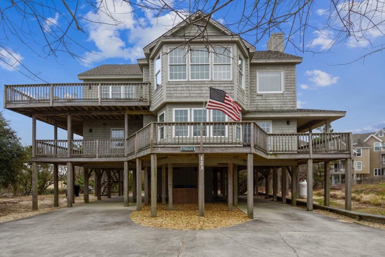 A distinctive coastal property elevated on sturdy pilings, featuring wraparound decks and classic shingle siding. The American flag marks the welcoming entrance to this beachside retreat, set among native coastal trees and just steps from other vacation h
