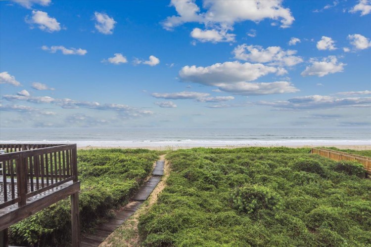 A wooden boardwalk leads through dune grass to the beach, with ocean and sky stretching endlessly ahead in the surrounding coastal landscape.