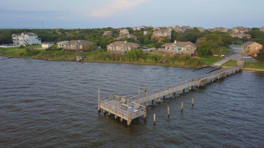 Aerial view of the waterfront community featuring a private fishing pier extending into calm waters, surrounded by coastal homes and lush vegetation in the background.