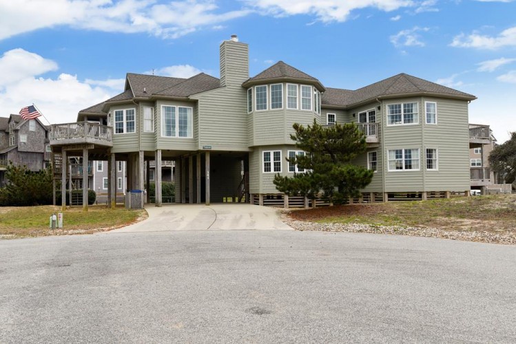 Expansive two-story coastal home on stilts with charming bay windows, multiple balconies, and ample parking in a peaceful beachside neighborhood.