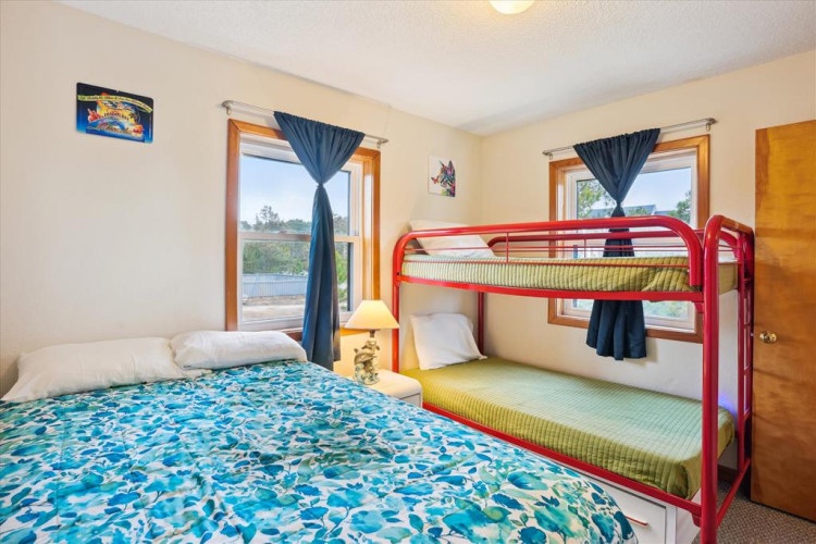 Your comfortable bedroom features a queen bed and a vibrant red bunk bed, perfect for families. Natural light fills the space through dual windows overlooking the water, while cozy touches await your arrival.