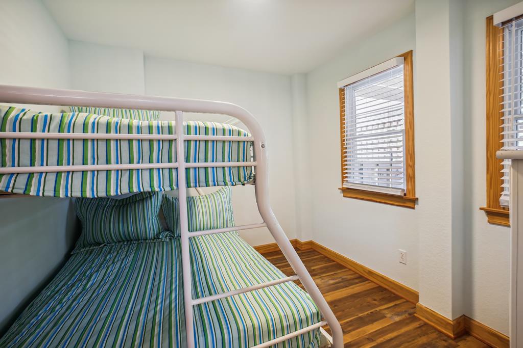 Bright bunk room with cheerful striped linens offers cozy sleeping space for kids or groups, featuring warm wood floors and natural light streaming through plantation shutters.