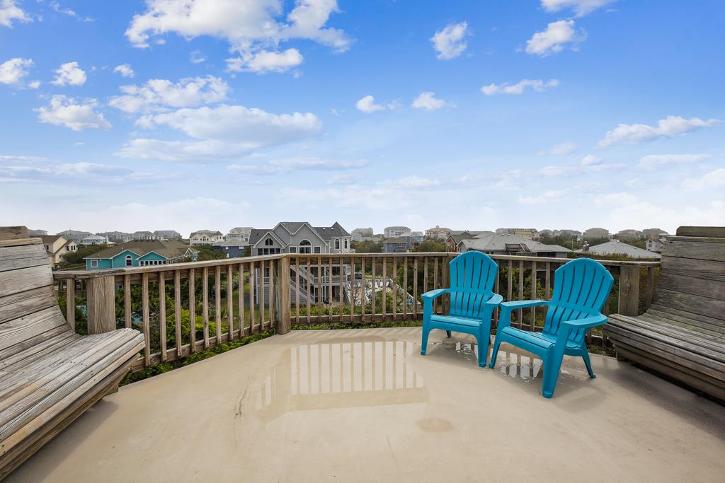 A rooftop deck overlooking a residential neighborhood with scenic views of surrounding homes and open skies. Two bright blue Adirondack chairs positioned for viewing the area.