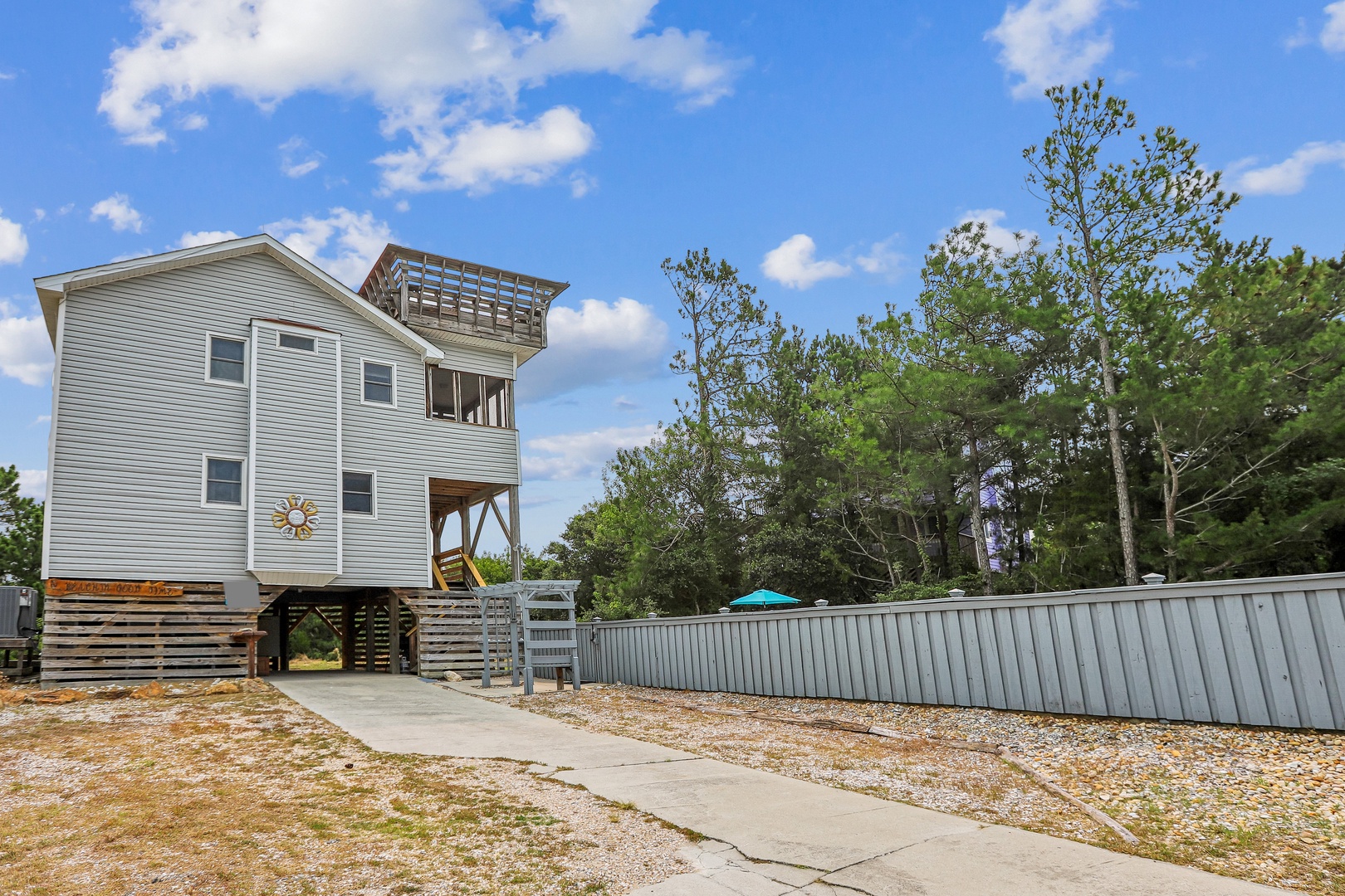 The property exterior features a distinctive observation deck, ground-level access, and parking areas, surrounded by mature coastal trees in a quiet residential setting.