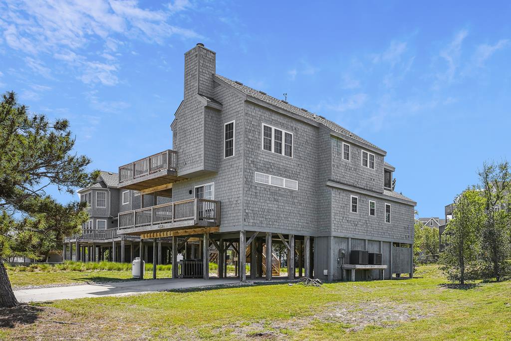 Gray-shingled coastal home on stilts with multilevel balconies, set in a grassy coastal area with mature trees and neighboring properties nearby.