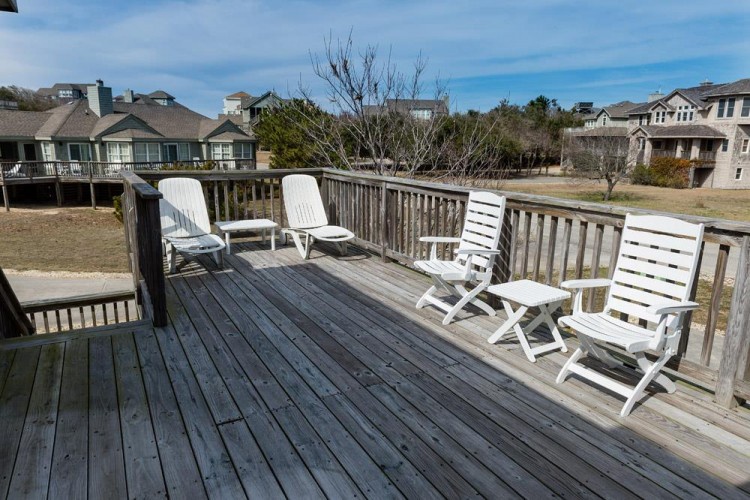 A spacious deck set in a residential coastal community, featuring white lounge chairs and surrounded by neighboring homes, offering an elevated outdoor space for relaxation.