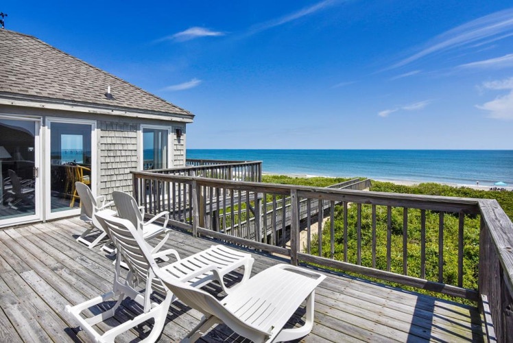 Relax on your private oceanfront deck, where white lounge chairs invite you to unwind while taking in sweeping views of turquoise waters and coastal greenery under endless blue skies.