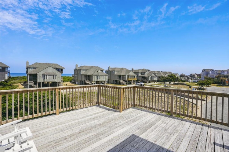 A spacious oceanfront deck offering panoramic views of the beach and neighboring coastal homes under clear blue skies, perfect for relaxing and soaking in the seaside setting.
