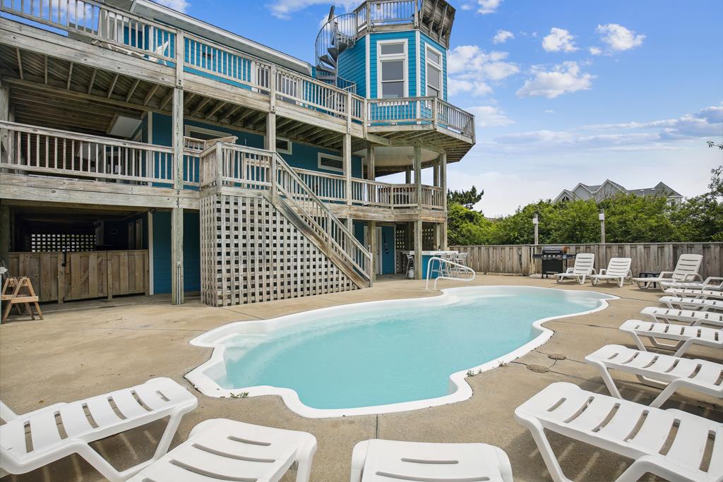 Exterior view shows a multi-level coastal rental property featuring a private pool surrounded by lounge chairs, multiple deck levels, and an observation tower, all set within a fenced yard in a residential beach community.
