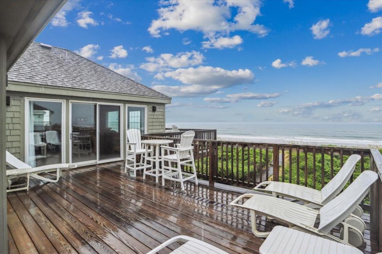 A stunning oceanfront deck awaits your arrival, with comfortable seating, direct beach access, and sweeping coastal views stretching to the horizon under endless blue skies.