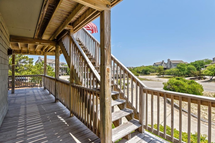 The property features an expansive covered deck with comfortable seating areas and glimpses of the surrounding coastal neighborhood through mature landscaping.