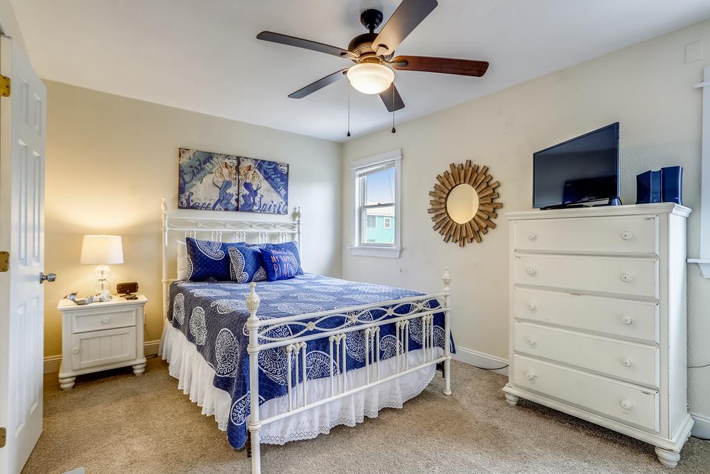 Your restful bedroom features a queen bed with coastal linens, elegant white furniture, a ceiling fan, flat-screen TV, and charming coastal décor that invites relaxation after a day at the beach.