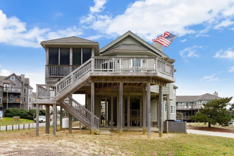 Coastal vacation home elevated on stilts, featuring screened porch and upper deck with American flag. Located in a quiet residential beach community.