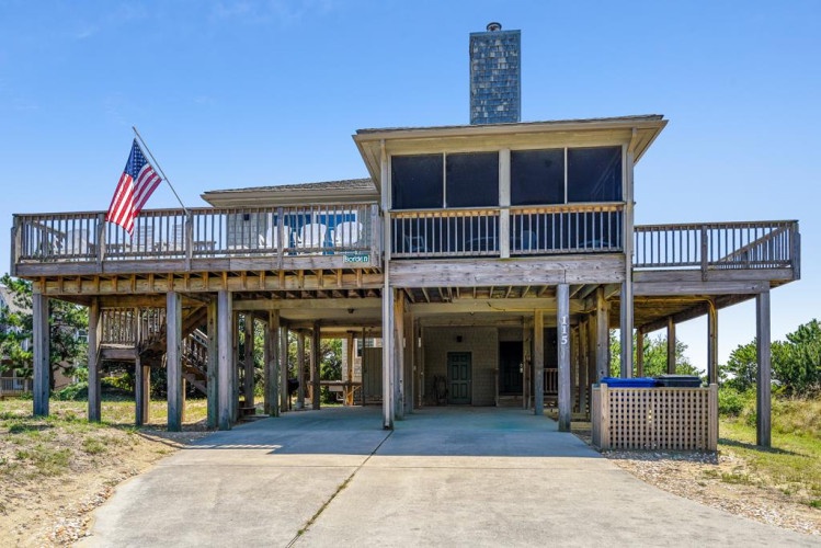 A coastal beach house elevated on stilts with wraparound decks, screened upper porch, and parking below—classic Outer Banks architecture designed for oceanfront living.